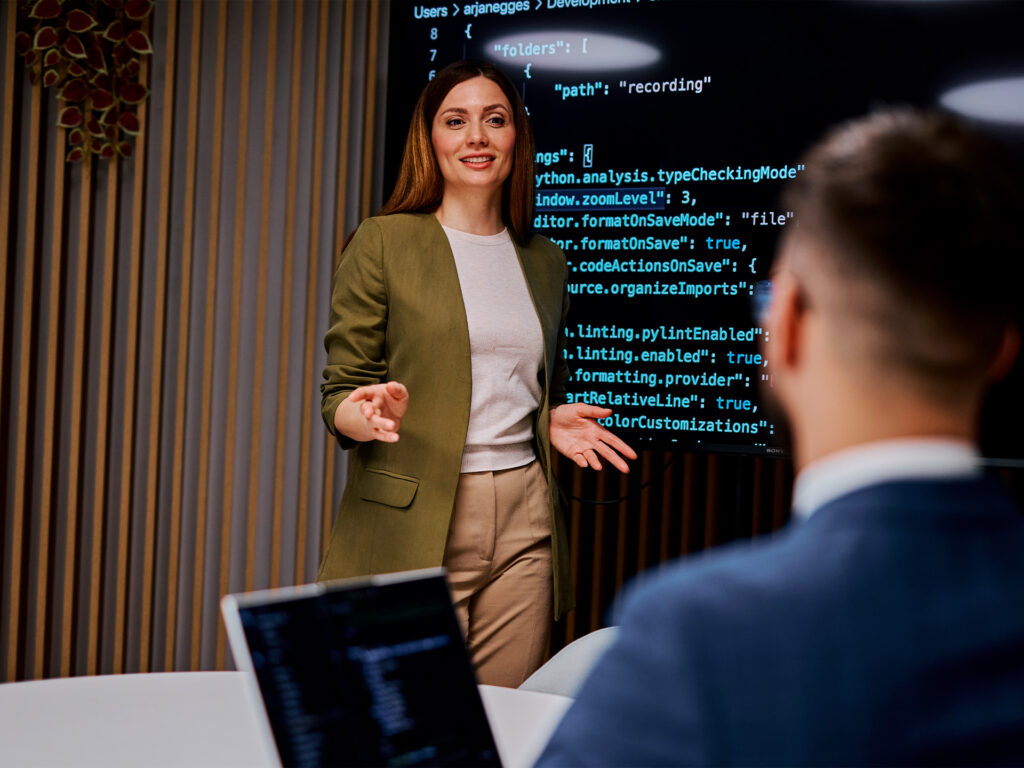 Person presenting code on screen to seated colleague in meeting room